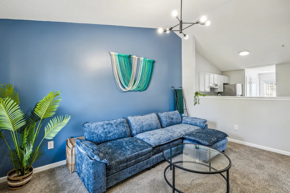 This image showcases a spacious living room with a blue sectional sofa, decorative wall hanging, indoor plant, and a glass coffee table. The room is filled with natural light and arranged to encourage both relaxation and socializing, with comfortable seating that invites guests to unwind.