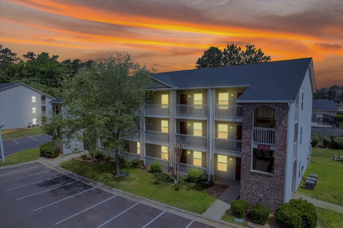 This image showcases the apartment building's exterior at sunset, including balconies, a parking area, and a warm facade. The golden hour lighting highlights the welcoming atmosphere and hints at the convenience of returning home after a day out.
