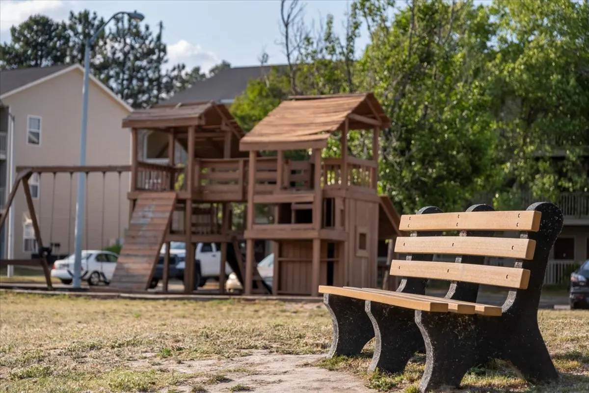An inviting outdoor playground area surrounded by green grass and trees, with a wooden bench nearby. The scene is lively, indicating opportunities for social gatherings, family activities, and relaxation within the community’s green spaces.
