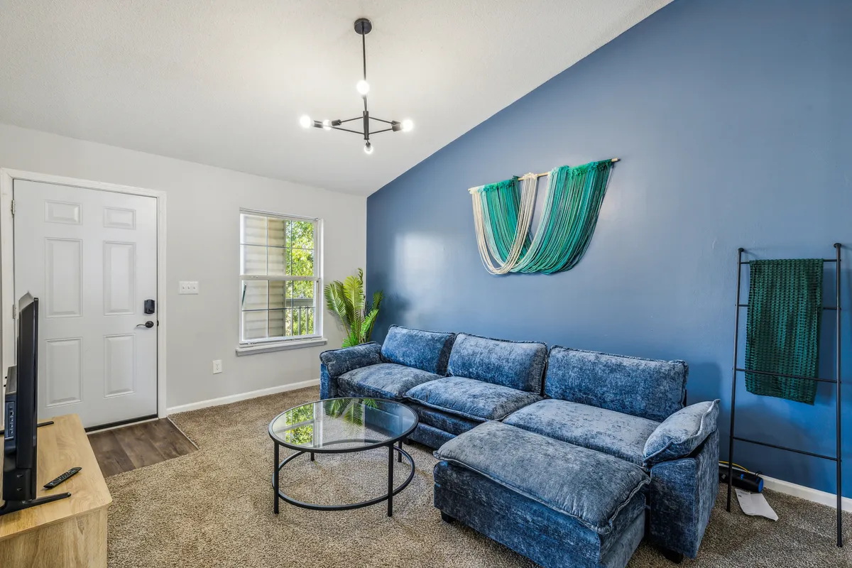 This image provides a close-up, cozy perspective of the living area, showcasing the sectional sofa, glass coffee table, textured wall hanging, green plant, and a modern light fixture. The natural light creates a warm, inviting atmosphere, highlighting the space's relaxing and social qualities.