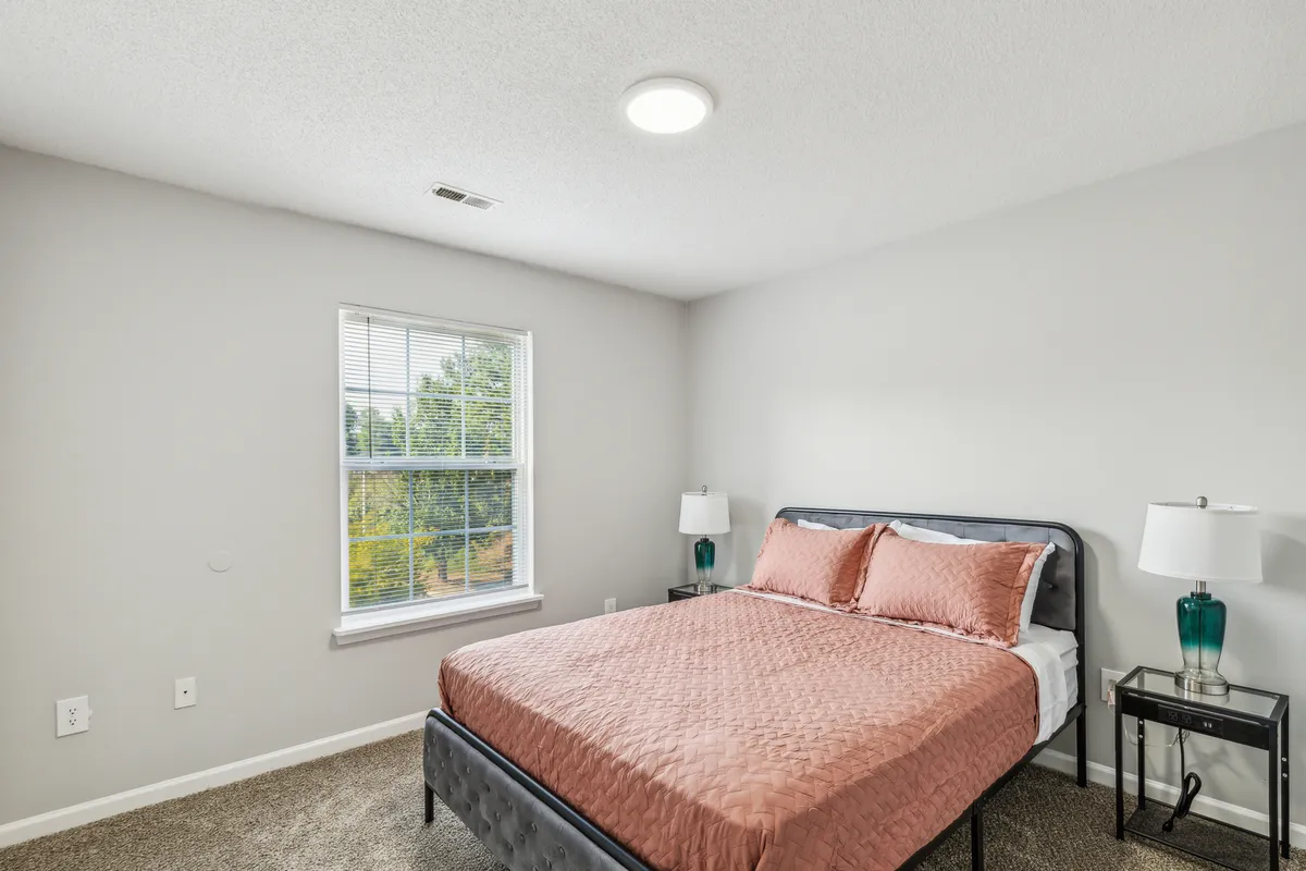 A serene bedroom awash in natural light, featuring a plush bed with inviting coral bedding, a stylish nightstand with a teal lamp, and a large window that fills the space with morning sunshine. The atmosphere is calm and inviting, perfect for moments of relaxation or reading in comfort.