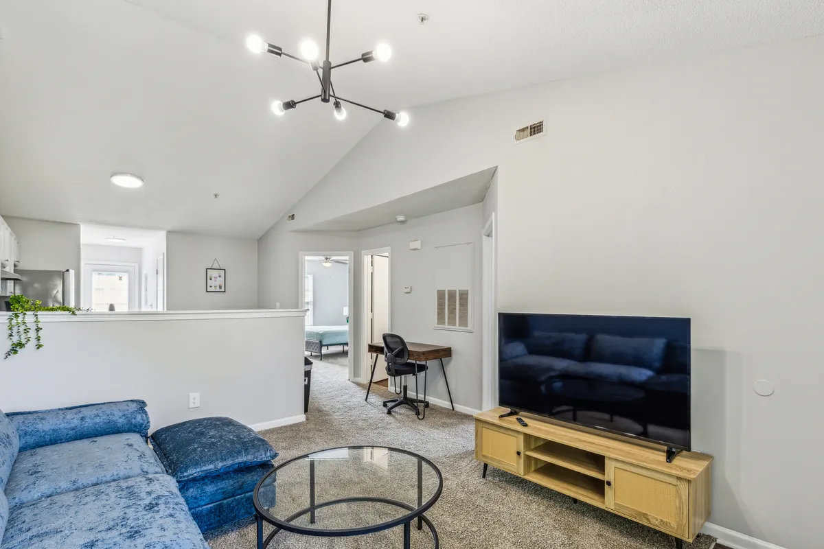 This scene presents another angle of the living area, featuring a blue sectional sofa, modern light fixture, flat-screen TV, and a glass coffee table. The room exudes a casual and comfortable vibe, perfect for movie nights or quiet mornings with coffee, as described in the section.