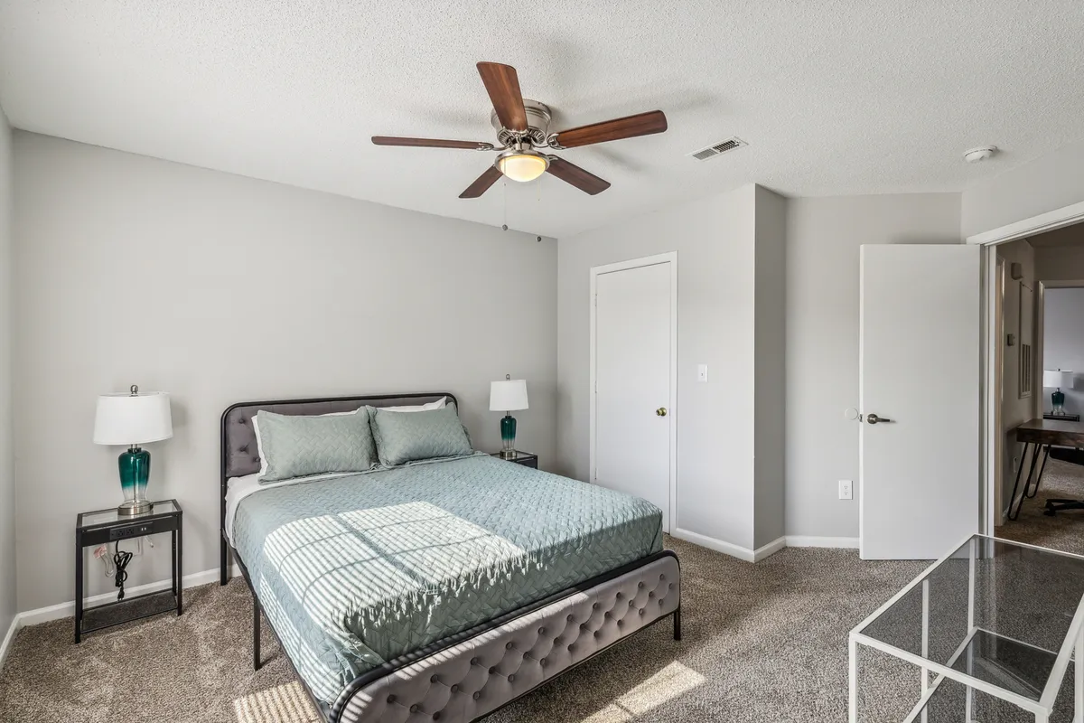 A tranquil bedroom featuring a plush queen bed with soft linens, a nightstand, a ceiling fan overhead, and a desk for work or reading. The room is filled with natural light, creating a relaxing and comfortable environment.