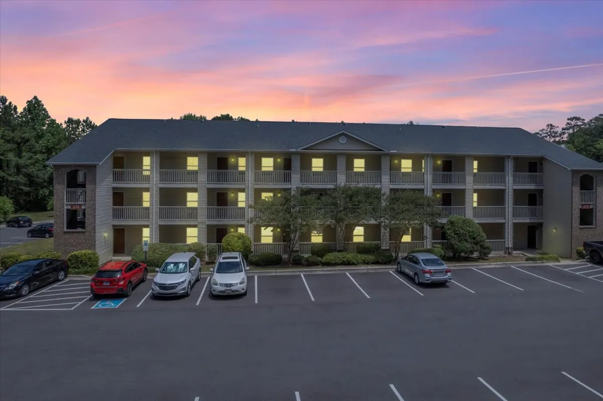 This image showcases the apartment building's exterior with a well-maintained parking area, attractive landscaping, and a dramatic sunset sky in the background. Balconies are visible, suggesting private outdoor spaces, and the overall mood is inviting and serene.